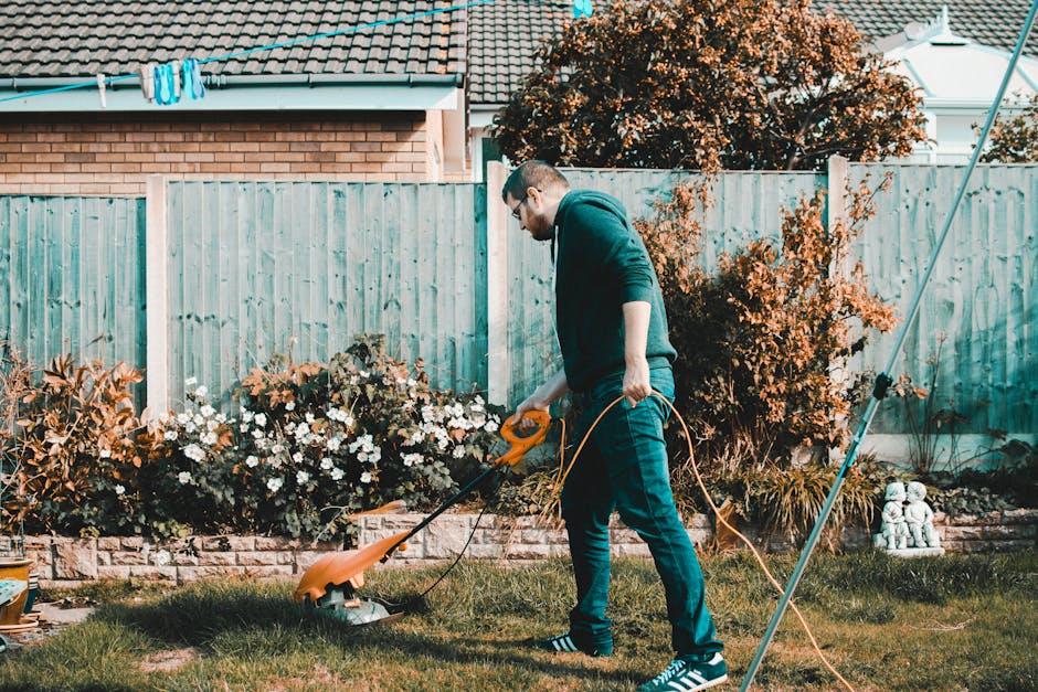 A man with glasses, wearing a dark hoodie and jeans, is operating an electric grass trimmer on a well-maintained lawn in a residential garden. The garden features a border of flowering plants, includi