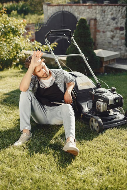 A man with glasses, wearing a dark hoodie and jeans, is operating an electric grass trimmer on a well-maintained lawn in a residential garden. The garden features a border of flowering plants, includi